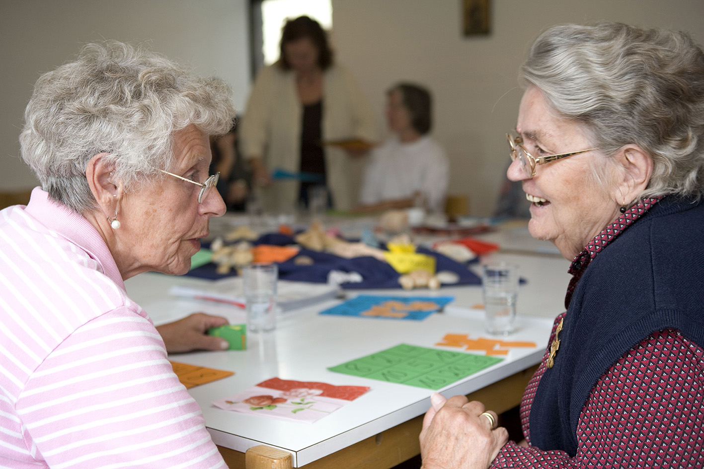 Zwei Seniorinnen sitzen an einem Tisch,auf dem verschiedene Bastelmaterialien liegen. Die Damen sind in ein Gespräch vertieft