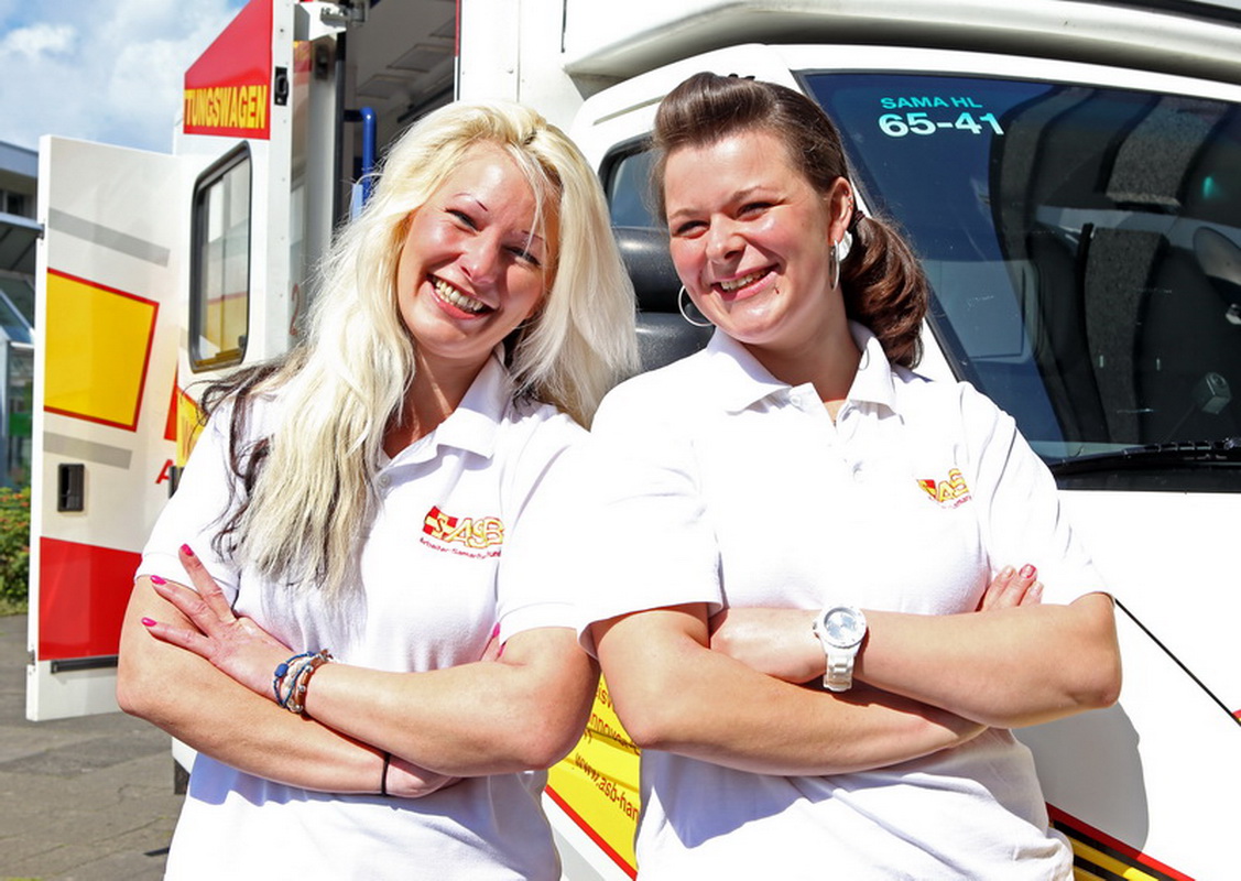 Zwei lächelnde Frauen mit verschränkten Armen lehnen an einem Fahrzeug der Samariter. Beide tragen ein weißes Poloshirt mit dem ASB-Logo